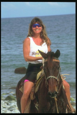 Женщина в очках прогуливается верхом на лошади по морском берегу