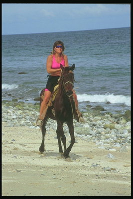 Женщина в красной футболке с удовольствием проезжает на лошади вдоль моря