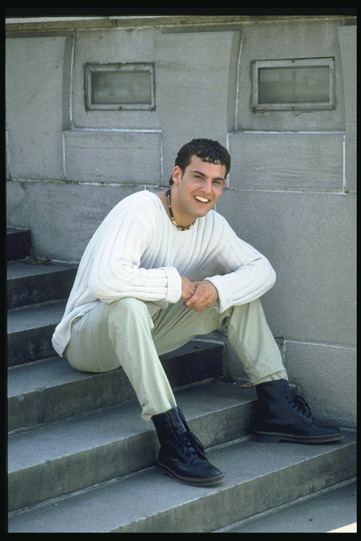 Vrolijke jonge man in een witte gebreide trui zitten op de trap