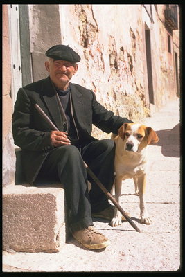 Vera. Një burrë ulet në hyrje të një shtëpi në një kostum të errët me një qen