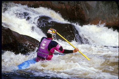 Ar zilu un dzeltenu kajaks un airis, sportists iet lejup pa upi