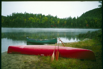 On the bank of the river is a red canoe with oars. Green canoe in the water near the shore
