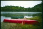 На березі річки лежить червоний каное з веслами. Зелений каное у воді біля берега